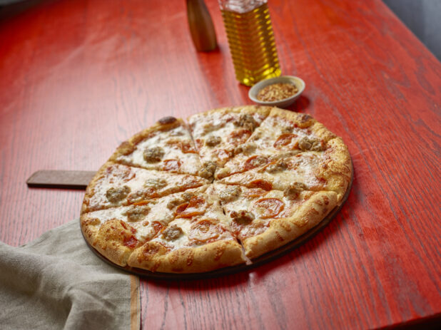 Sliced medium pizza with pepperoni and sausage with toppings in the background on a red wooden table