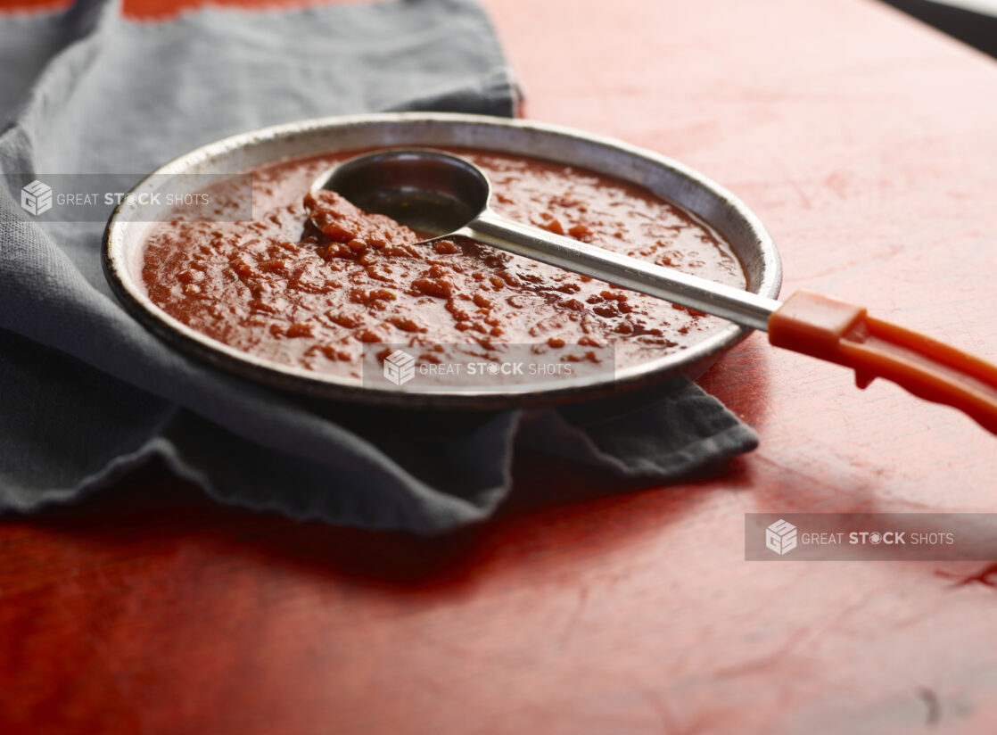 Shallow bowl of pizza sauce on a red wooden table with ladle scooping sauce, close-up