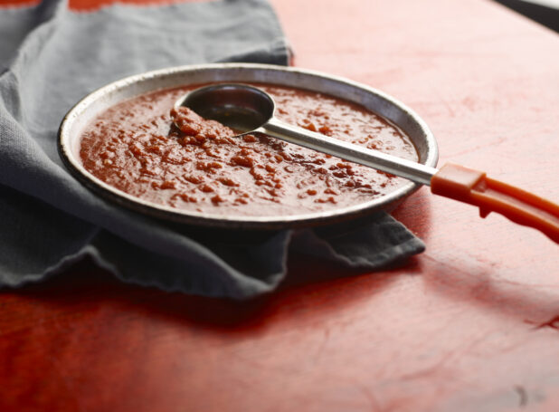 Shallow bowl of pizza sauce on a red wooden table with ladle scooping sauce, close-up