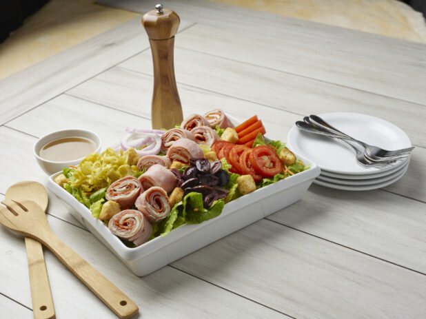 Large salad with rolled meats and a side dressing with servewear in the background