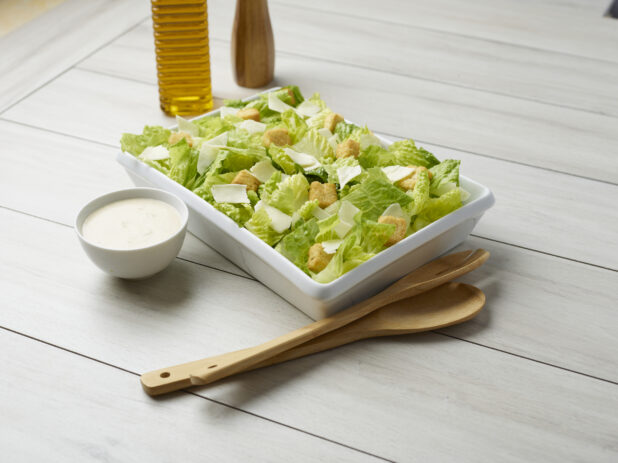 Large Caesar salad with dressing on the side and servewear in the background