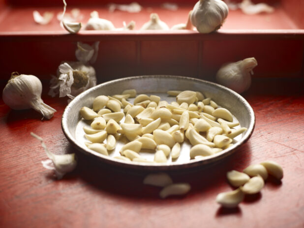Shallow metal pan filled with garlic cloves on a red wooden background surrounded by garlic bulbs and more garlic cloves