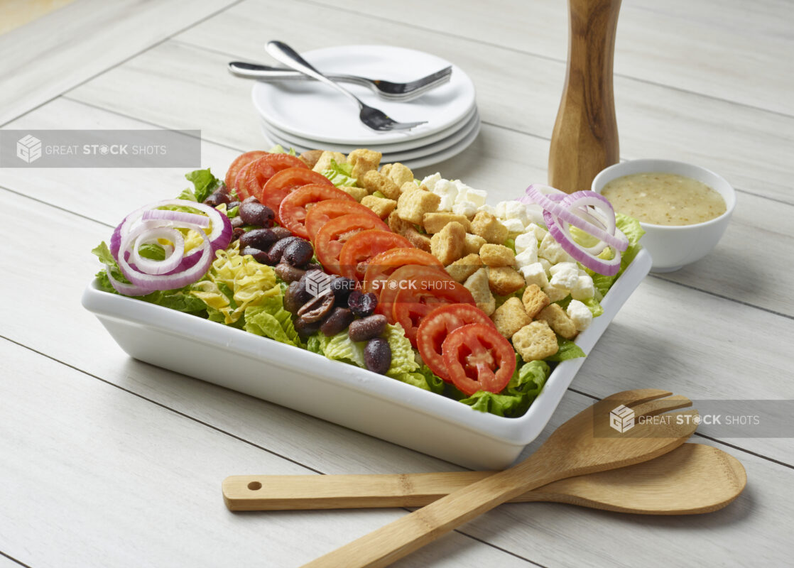 Large Greek salad with dressing on the side and servewear in the background