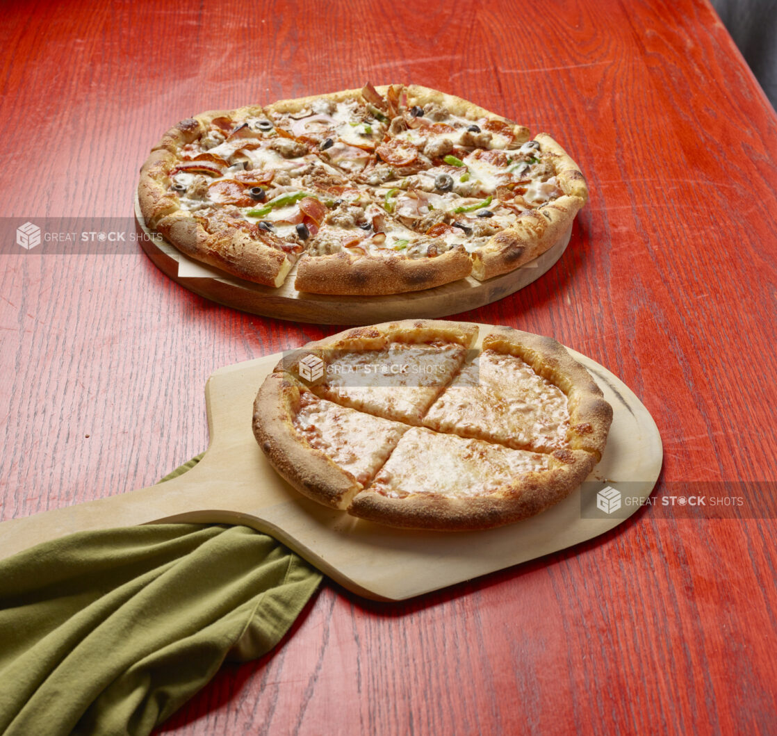 Two sliced pizzas, one medium with five toppings and one small with cheese on a red wooden table