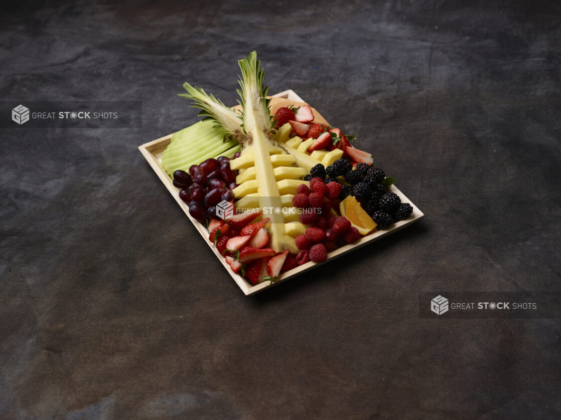 Assorted fruits on a wood tray with pineapple centerpiece on a dark background