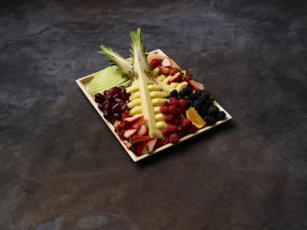 Assorted fruits on a wood tray with pineapple centerpiece on a dark background