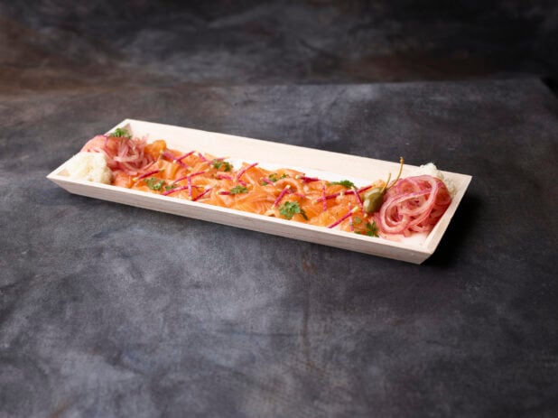 Thinly sliced smoke trout with pickled red onion and garnish on a wooden platter with a dark background