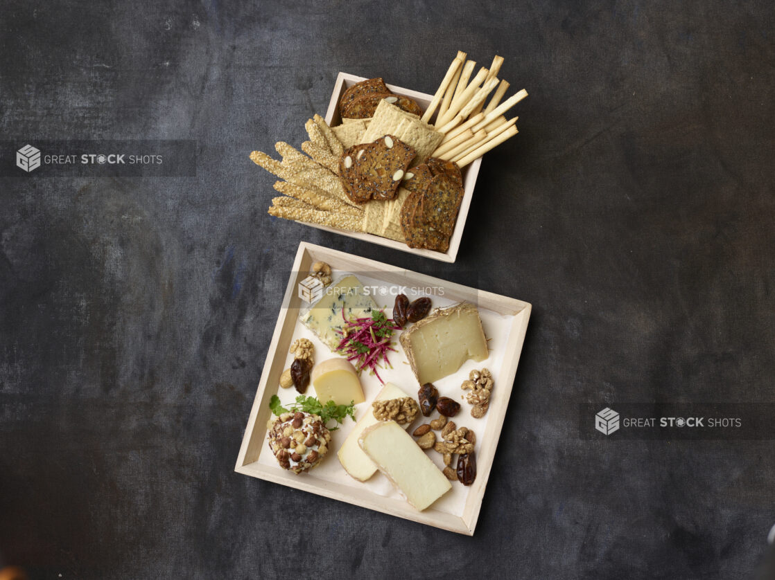 Overhead of assorted cheese and nuts on a wooden tray with a wooden bowl of breadsticks and crackers on a dark background