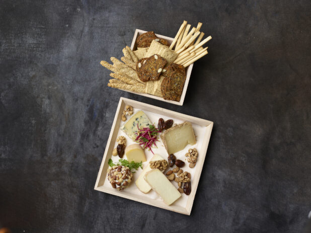 Overhead of assorted cheese and nuts on a wooden tray with a wooden bowl of breadsticks and crackers on a dark background