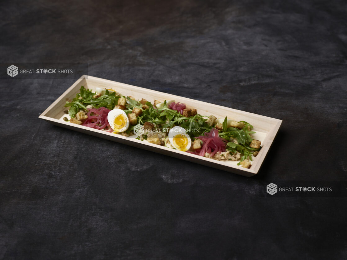 Fresh tuna and arugula salad with pickled red onions, eggs, croutons and capers in a wooden tray on a dark background