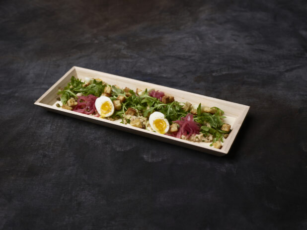 Fresh tuna and arugula salad with pickled red onions, eggs, croutons and capers in a wooden tray on a dark background