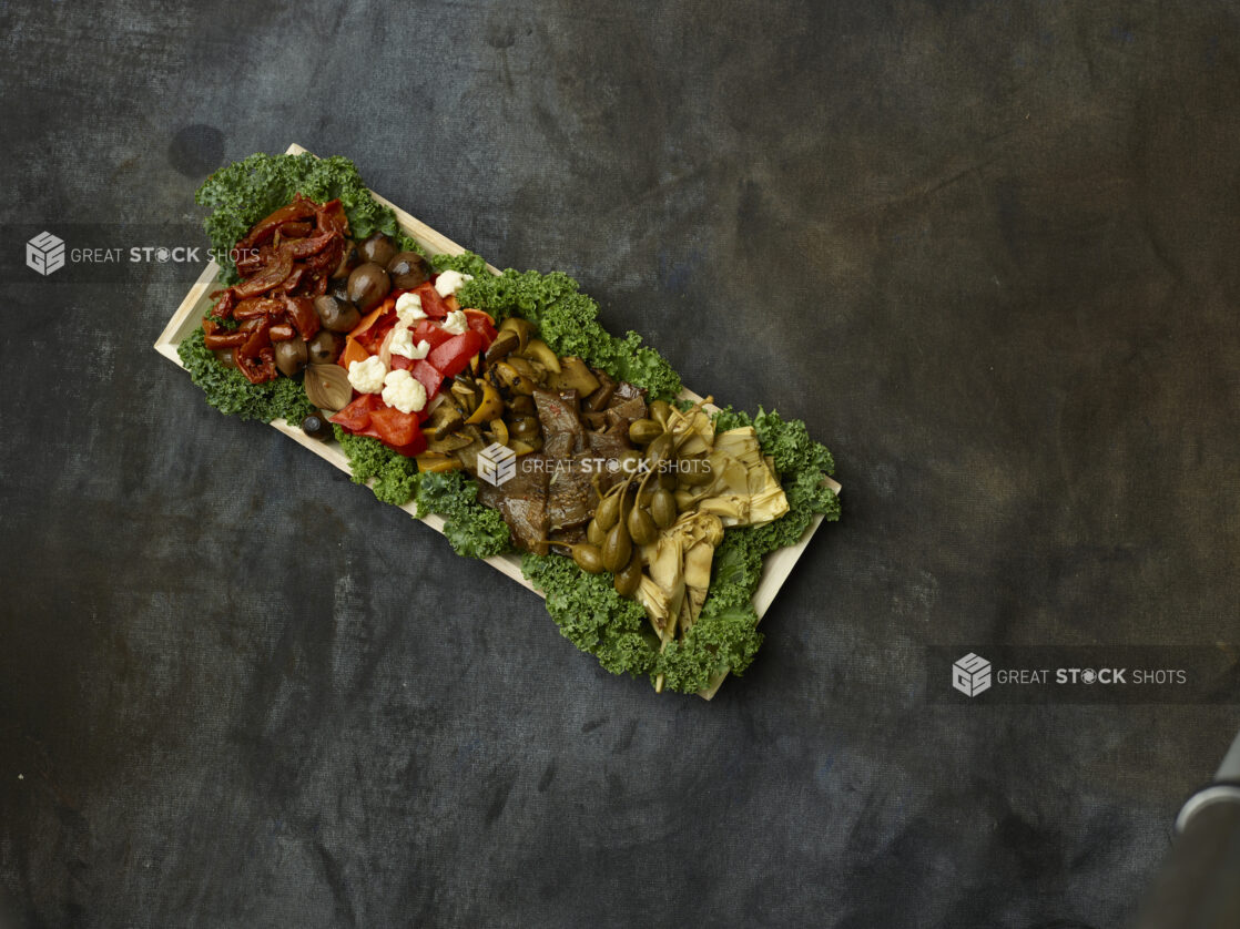 Overhead View of an Antipasto Catering Tray with Assorted Pickled, Marinated, Roasted and Grilled Vegetables on a Bed of Fresh Kale on a Black Canvas Surface