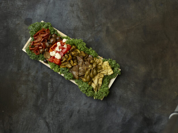 Overhead View of an Antipasto Catering Tray with Assorted Pickled, Marinated, Roasted and Grilled Vegetables on a Bed of Fresh Kale on a Black Canvas Surface