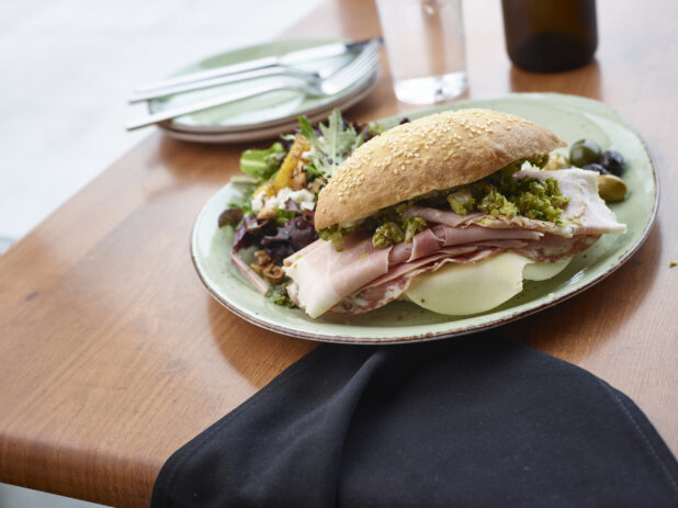 Italian Muffuletta Sandwich with a Side of Roasted Corn, Red Beets and Arugula Salad and Marinated Olives on a Green Ceramic Dish in a Restaurant Setting