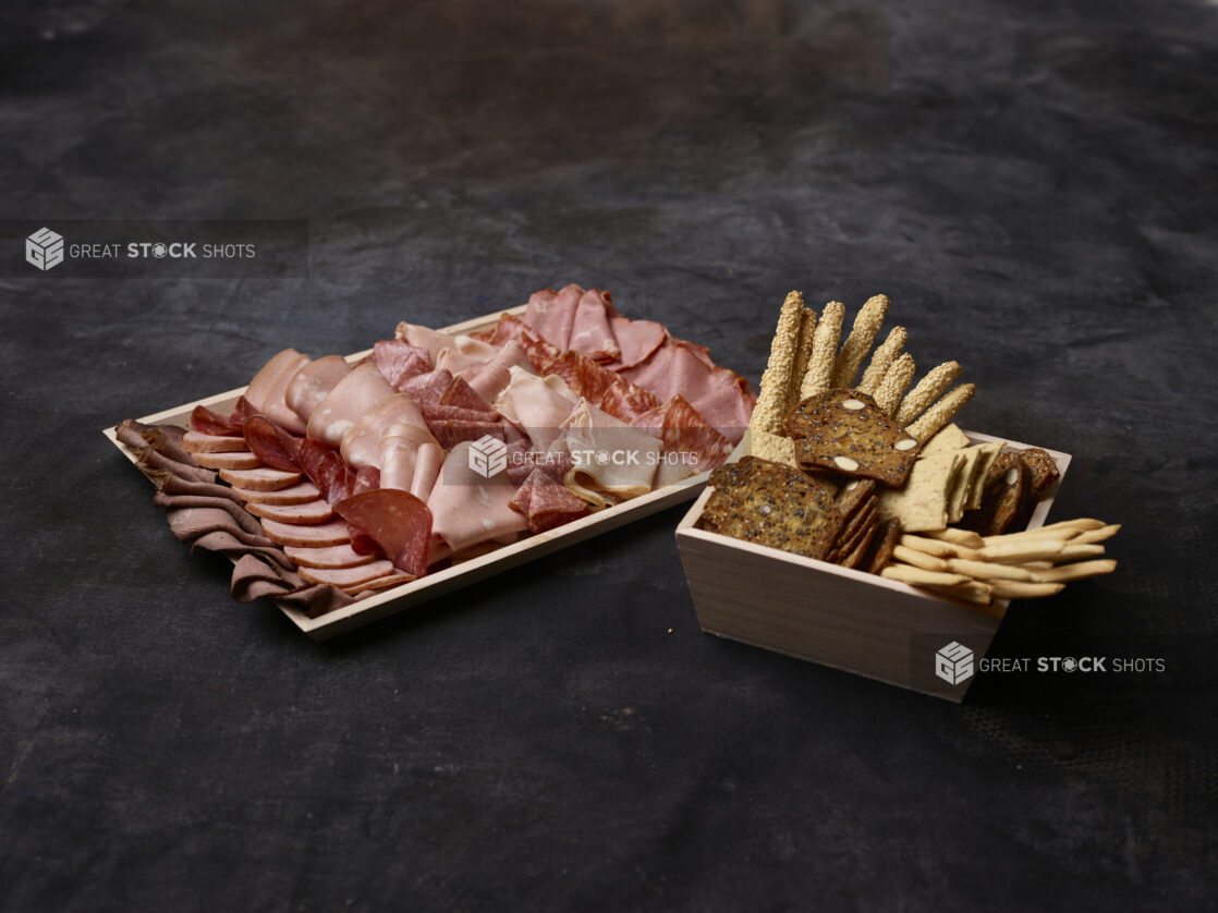 Wooden platter of cured sliced deli meats with a wooden bowl of assorted crackers and breadsticks