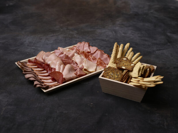 Wooden platter of cured sliced deli meats with a wooden bowl of assorted crackers and breadsticks