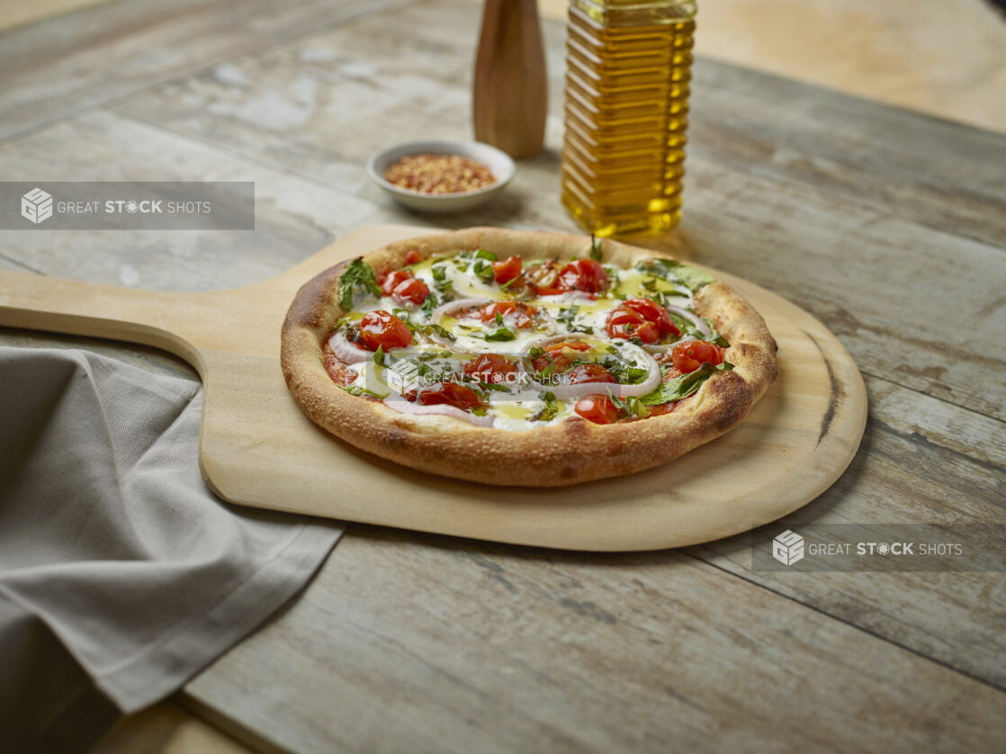 Whole pizza with cherry tomatoes, onion and fresh basil on a paddle with toppings in the background