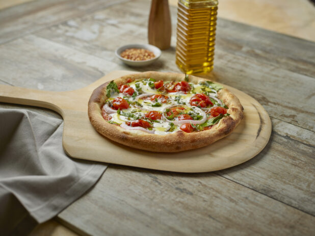 Whole pizza with cherry tomatoes, onion and fresh basil on a paddle with toppings in the background