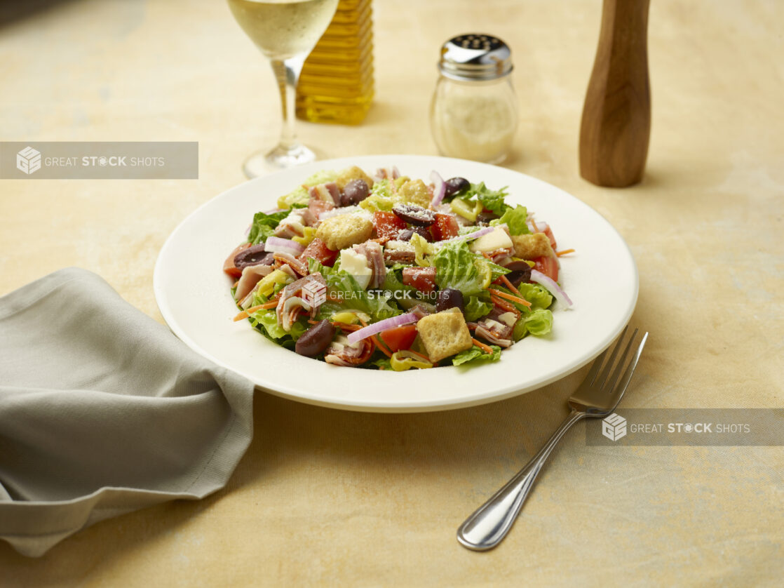 Salad with sliced ham, salami and provolone squares on top of red onion, black olives and croutons with a glass of white wine on a beige background