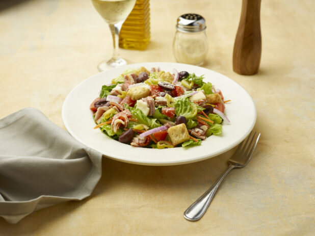 Salad with sliced ham, salami and provolone squares on top of red onion, black olives and croutons with a glass of white wine on a beige background
