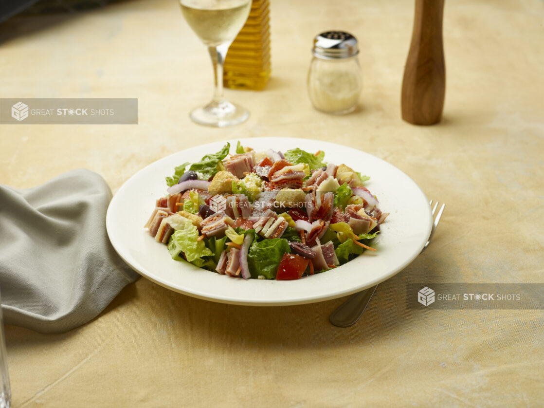 Salad with sliced ham, salami and provolone squares on top of red onion, black olives and croutons with a glass of white wine on a beige background