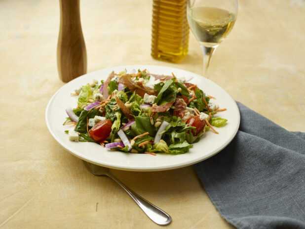 Salad with bacon, blistered cherry tomatoes, red onion, blue cheese and pine nuts with a glass of white wine on a beige background