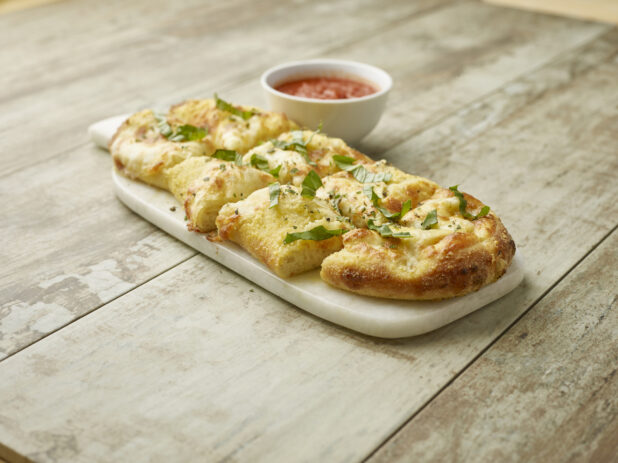 Cut garlic cheese bread topped with fresh basil on a marble board with a side of tomato sauce