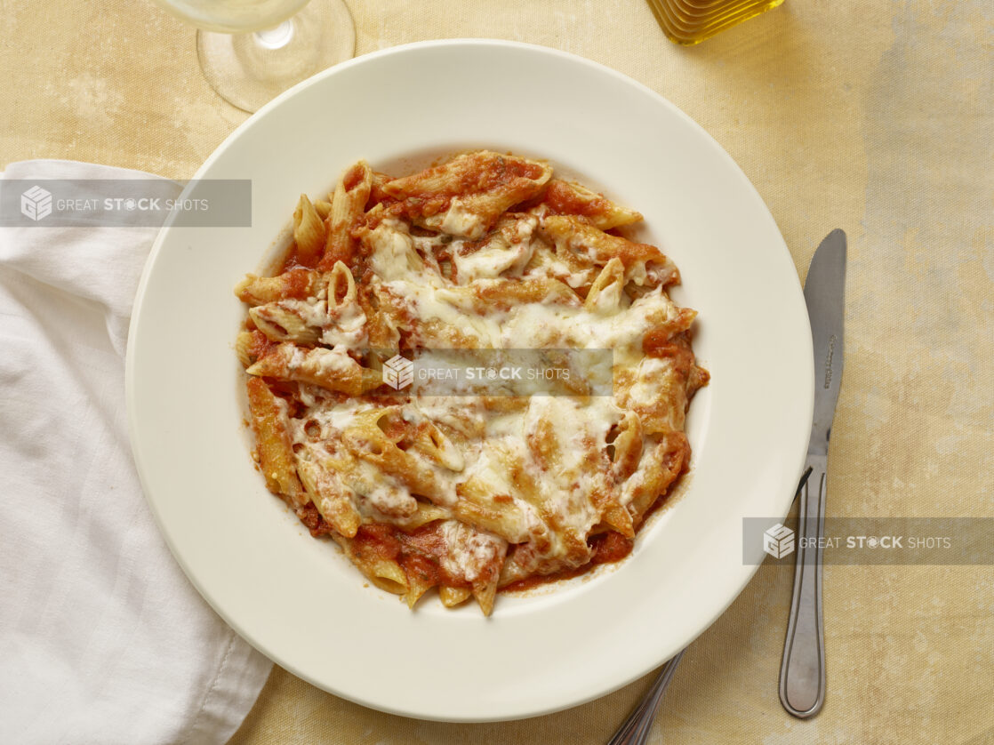 Overhead bowl of penne pasta in tomato sauce with melted cheese on a yellow background