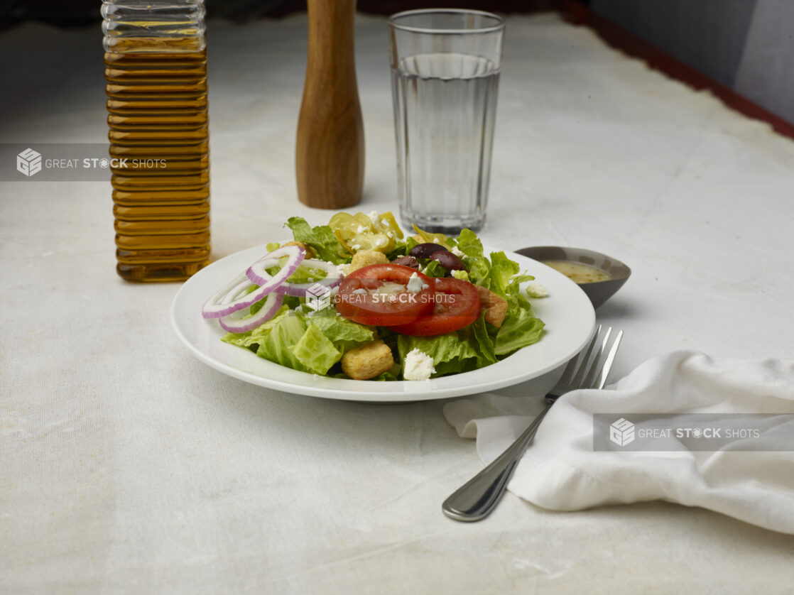 Greek salad with dressing on the side on a white table cloth with glass of water in the background