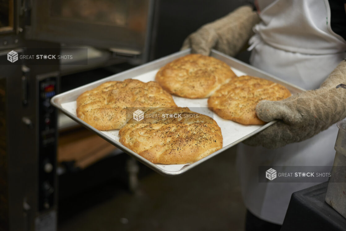 Close Up of an Employee with Oven Mitts Holding a Baking Sheet of Fresh Baked Mini Herbed Focaccia Bread in the Kitchen of a Gourmet Grocery Store