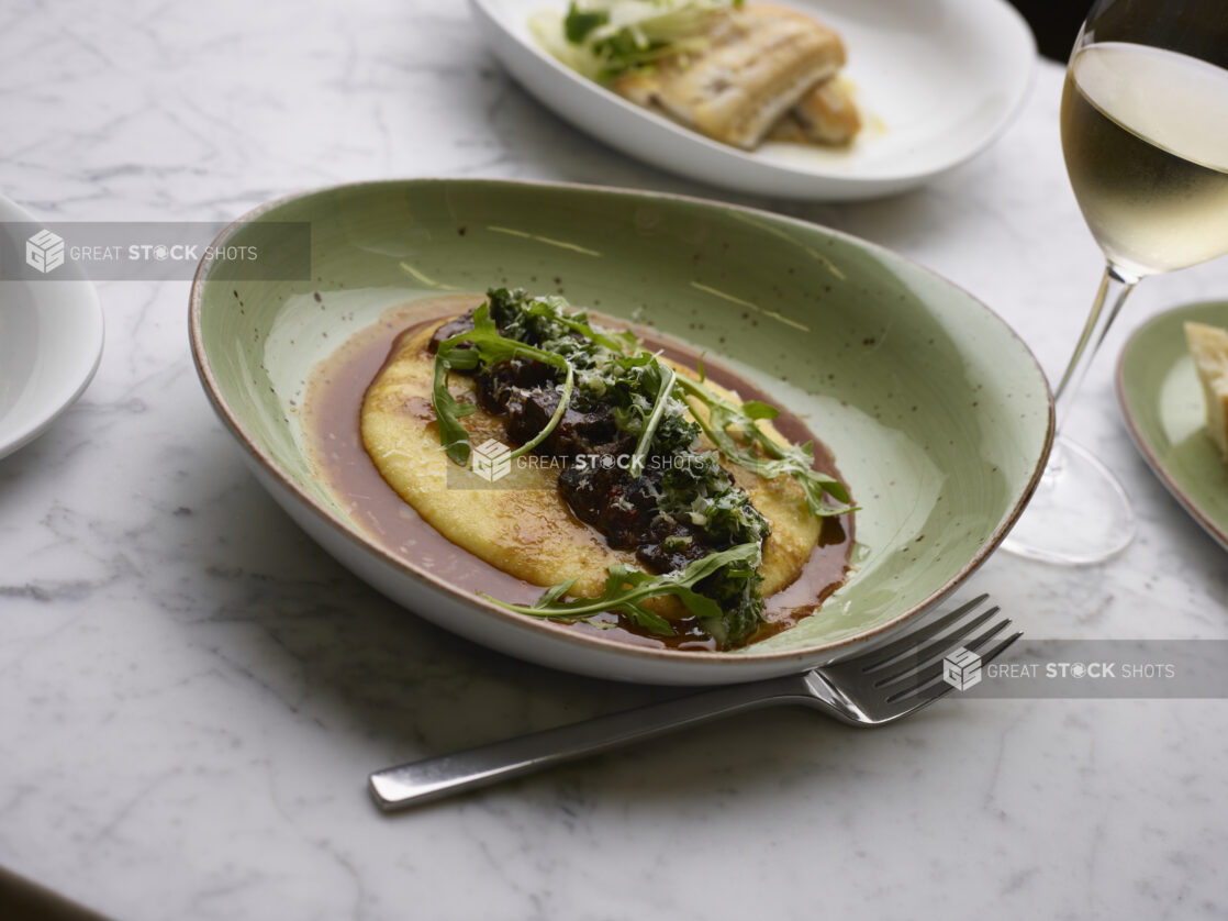 Braised Beef Short Ribs and Baby Arugula on a Bed of Polenta with Au Jus in a Green Ceramic Dish on a Marble Table - Variation