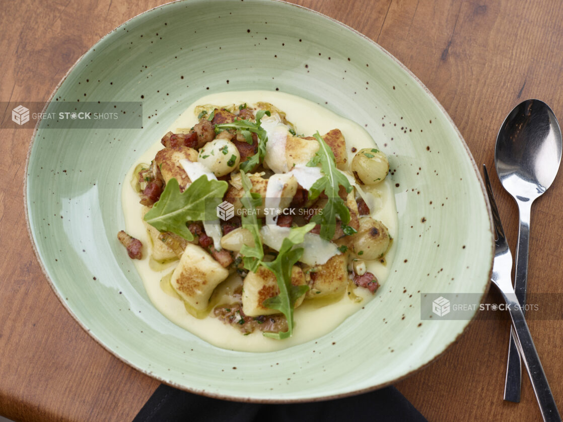 Overhead of large green bowl of creamy gnocchi and bacon garnished with arugula and shaved parmesan cheese