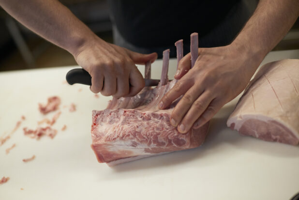Butcher's hands cutting pork chops with long bones on a white cutting board, close-up