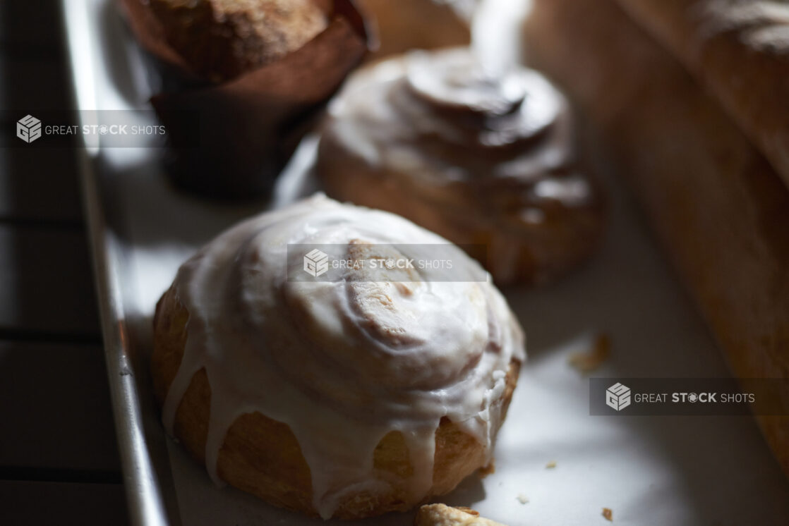 Close Up of Fresh Baked Cinnamon Rolls with Melted Cream Cheese Frosting on a Baking Sheet with other Baked Goods in a Gourmet Grocery Store Setting