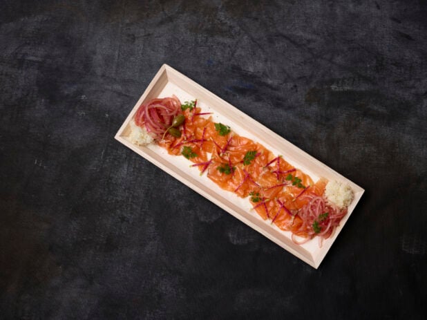 Overhead of thinly sliced smoked trout with pickled red onion and garnish on a wooden platter with a dark background