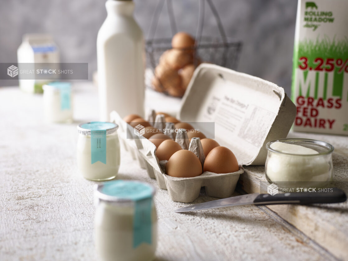 A carton of brown eggs surrounded by assorted dairy products on a weathered wood surface, close-up