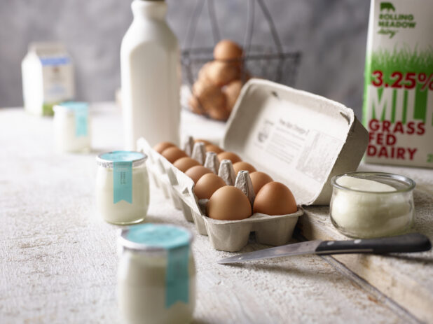 A carton of brown eggs surrounded by assorted dairy products on a weathered wood surface, close-up