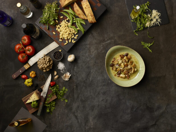 Bowl of gnocchi and many fresh ingredients on a dark grey cloth background