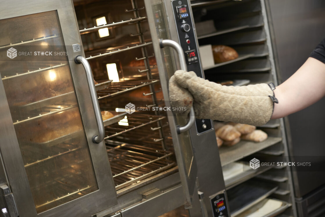 Oven Mitt-Covered Hand of an Employee Opening the Doors of a Commercial Baking Oven in the Bakery Kitchen of a Gourmet Grocery Store