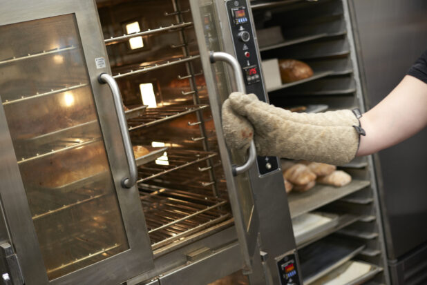 Oven Mitt-Covered Hand of an Employee Opening the Doors of a Commercial Baking Oven in the Bakery Kitchen of a Gourmet Grocery Store