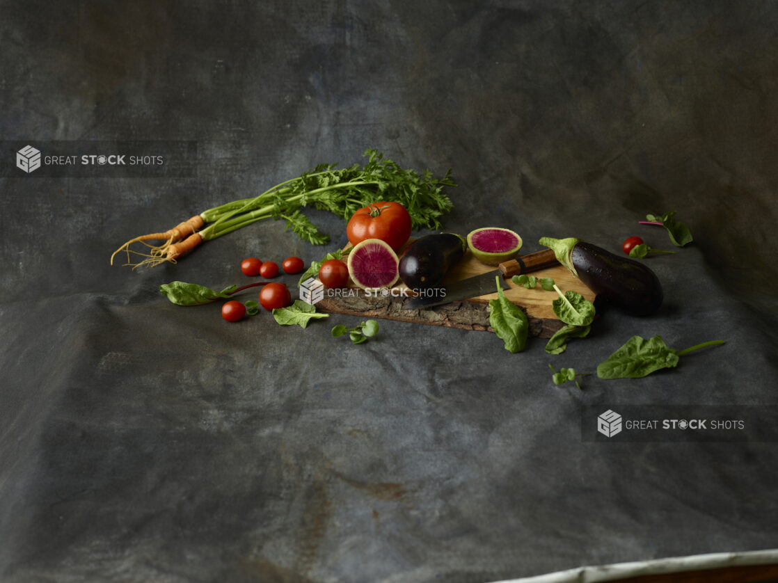 Collection of vegetables, whole and cut, on a live edge wooden board with a knife on a dark background