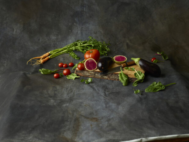 Collection of vegetables, whole and cut, on a live edge wooden board with a knife on a dark background