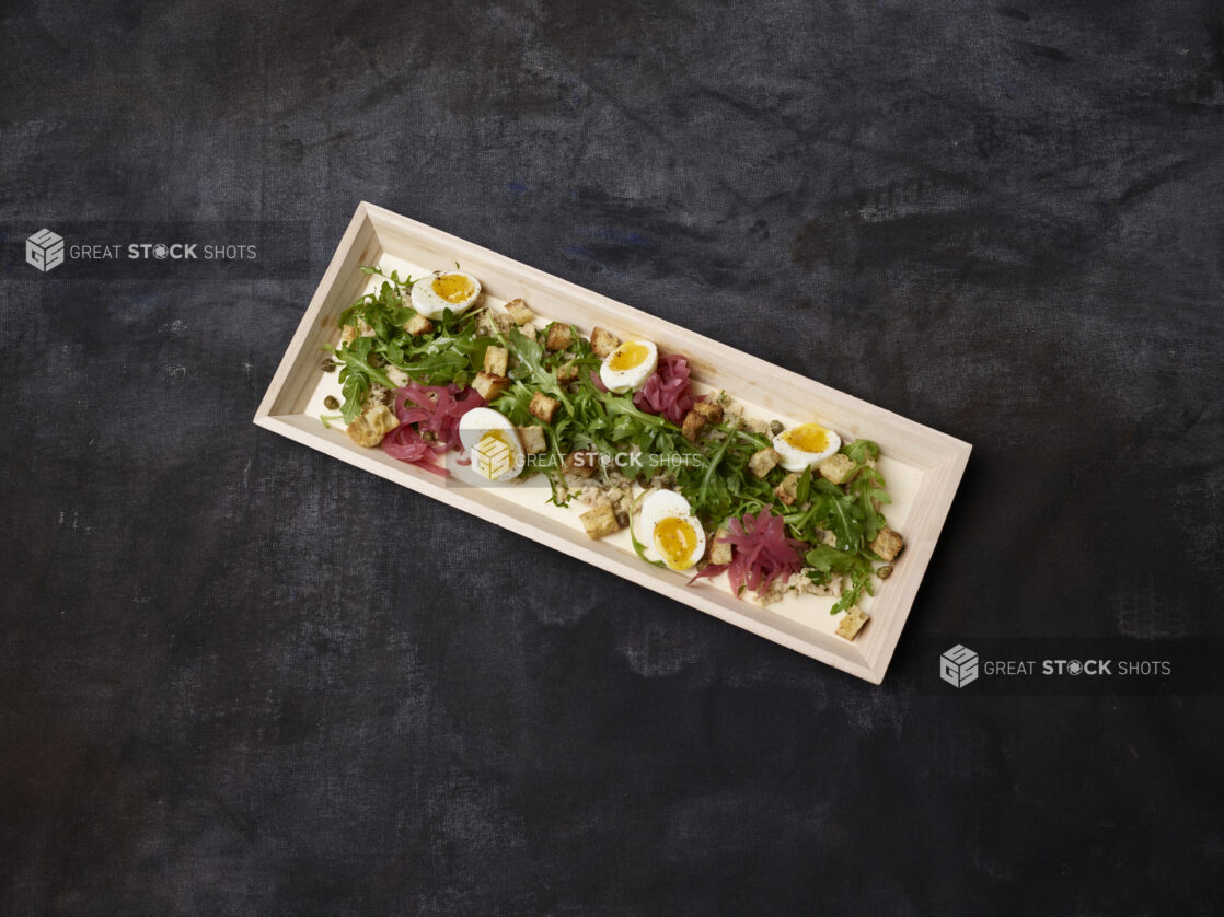 Overhead of fresh tuna and arugula salad with pickled red onions, eggs, croutons and capers in a wooden tray on a dark background