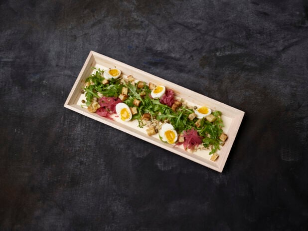 Overhead of fresh tuna and arugula salad with pickled red onions, eggs, croutons and capers in a wooden tray on a dark background