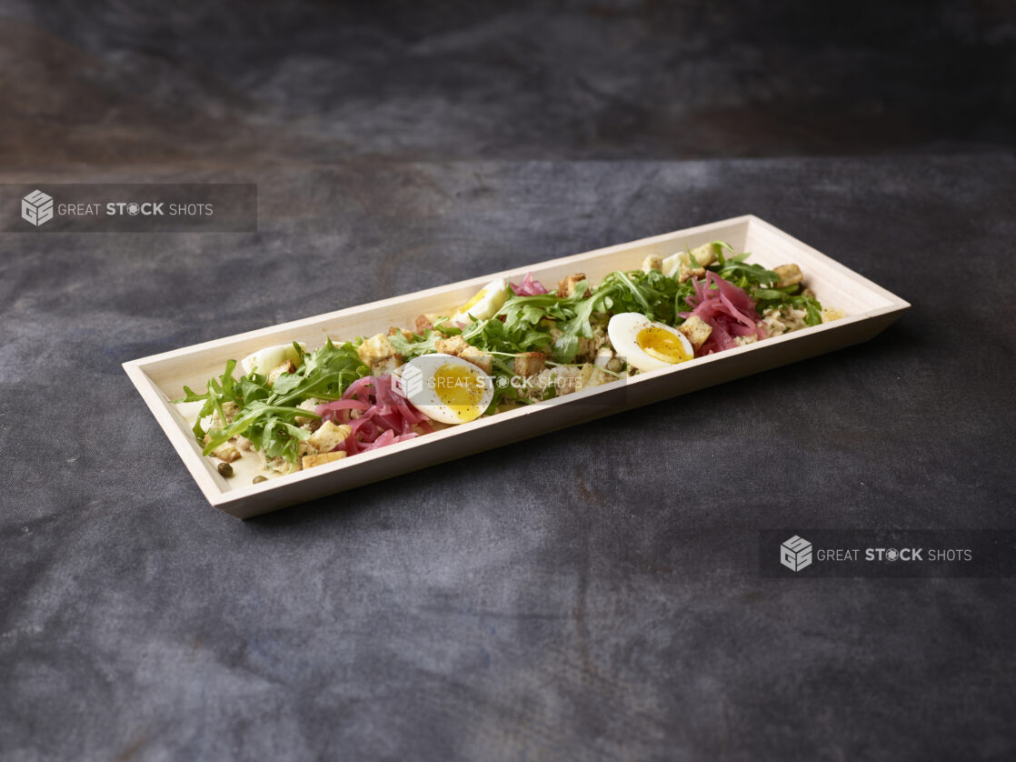 Fresh tuna and arugula salad with pickled red onions, eggs, croutons and capers in a wooden tray on a dark background