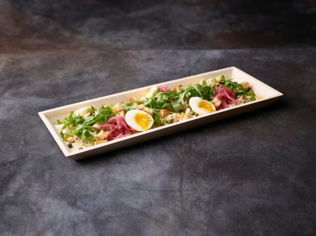 Fresh tuna and arugula salad with pickled red onions, eggs, croutons and capers in a wooden tray on a dark background