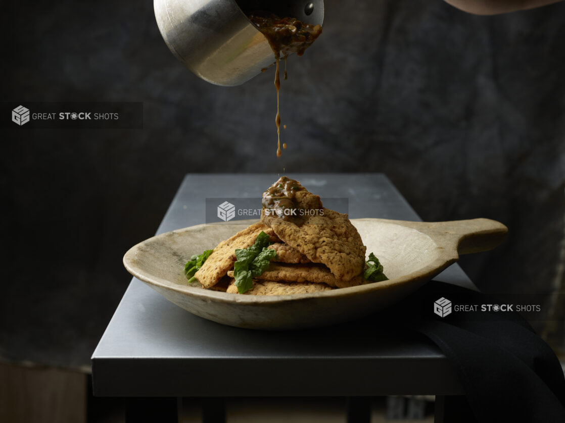 Wooden bowl of stacked fried chicken with fresh basil being drizzled with a brown marsala sauce