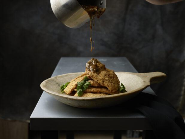 Wooden bowl of stacked fried chicken with fresh basil being drizzled with a brown marsala sauce