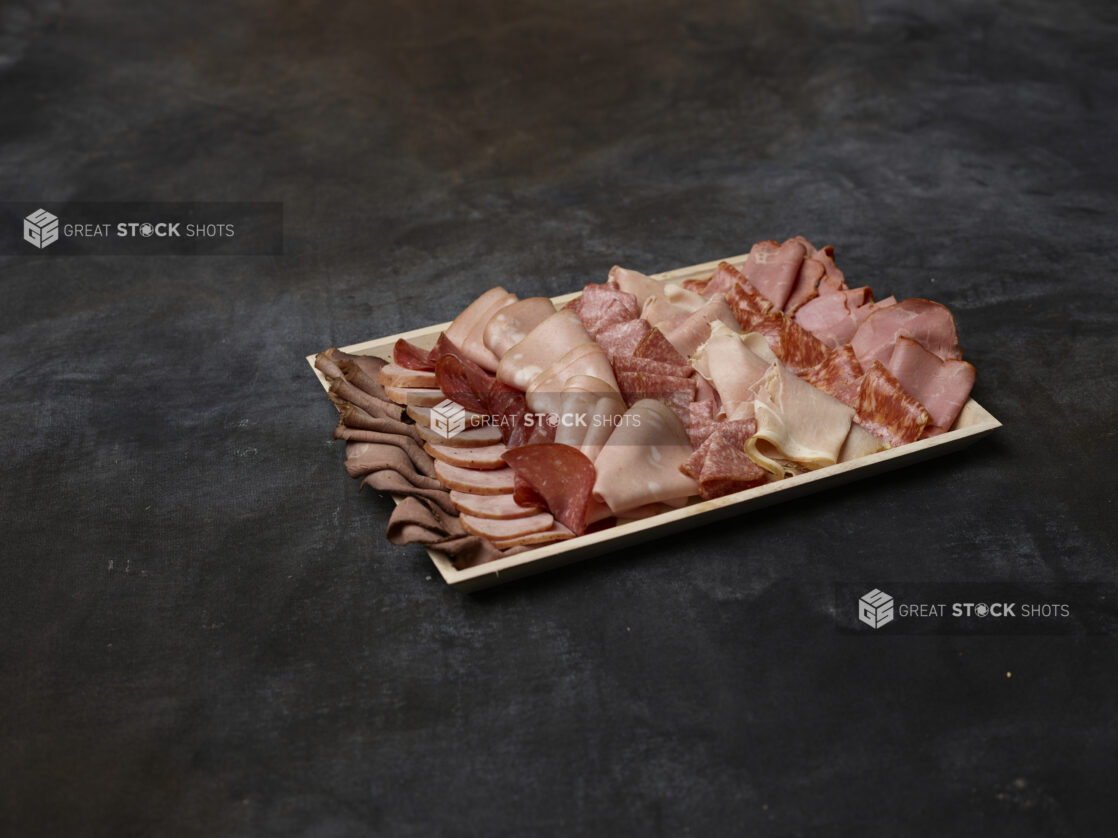 Wooden platter of cured sliced deli meats on a dark background