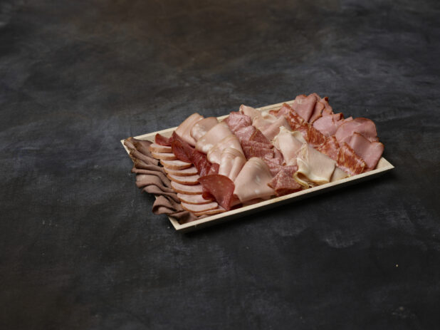 Wooden platter of cured sliced deli meats on a dark background